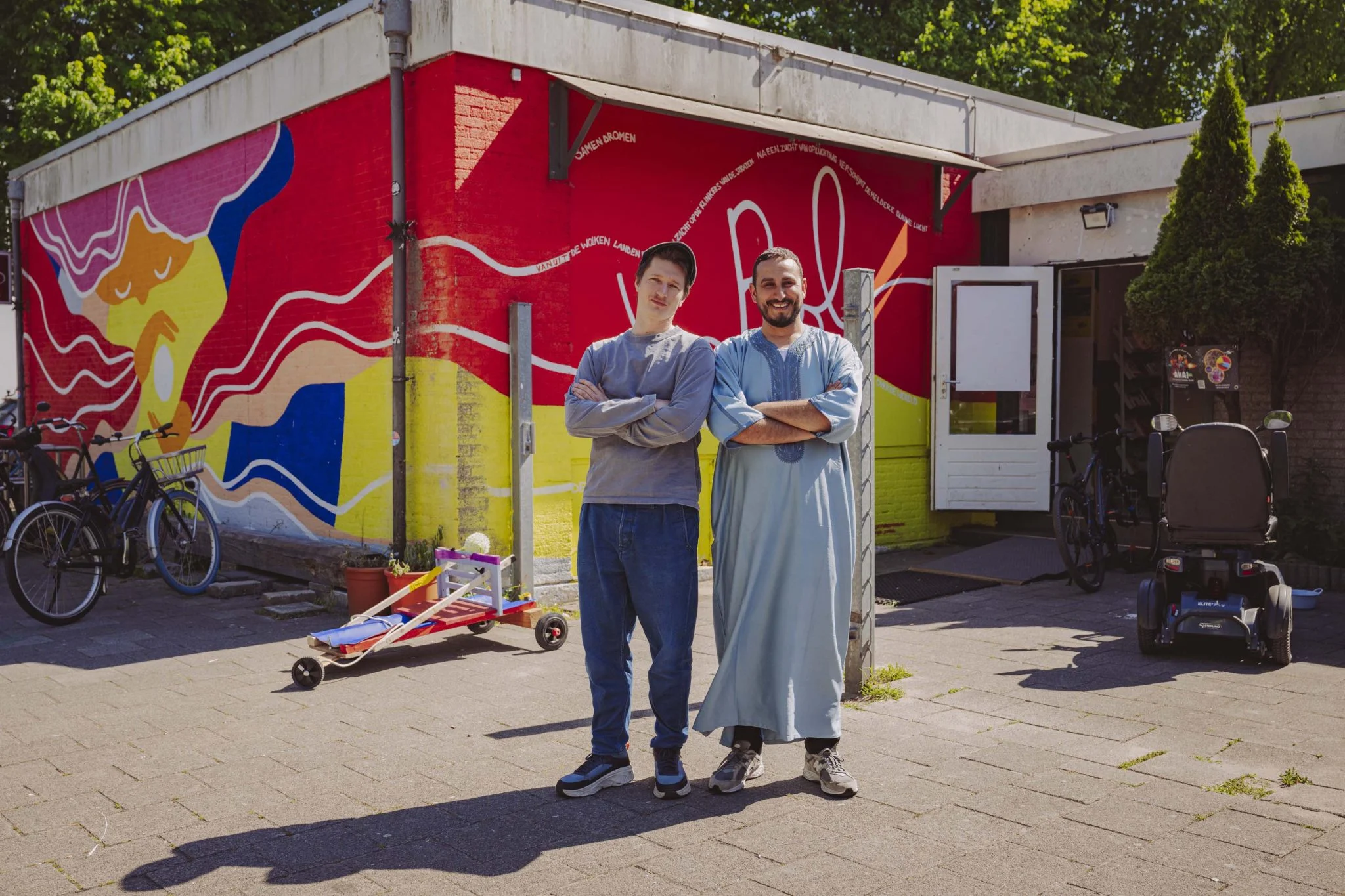 Two initiators in front of a colourful mural in the neighbourhood, with bikes and go-karts in front of it