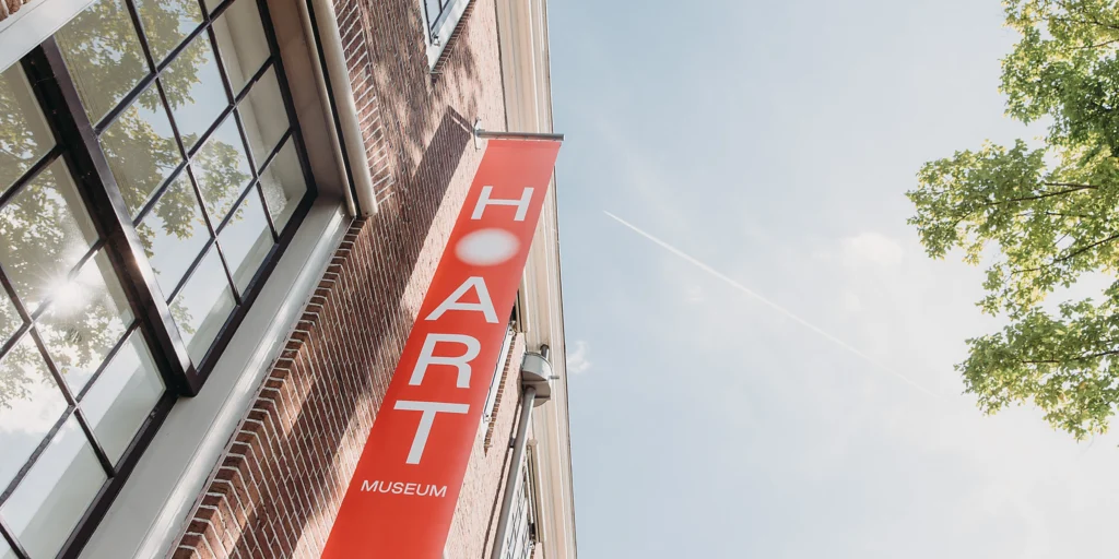 A red banner with the H’ART Museum logo hanging on the brick facade of the building on the Amstel