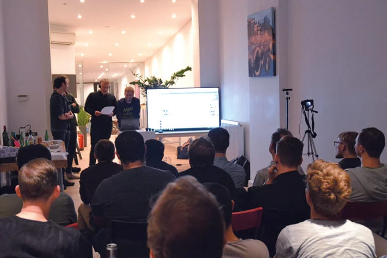 Een volle zaal bij een kennissessie op kantoor, met twee sprekers en een presentatie op het scherm